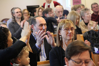 People in pews applauding, focused on one man in the middle with a kippah. 