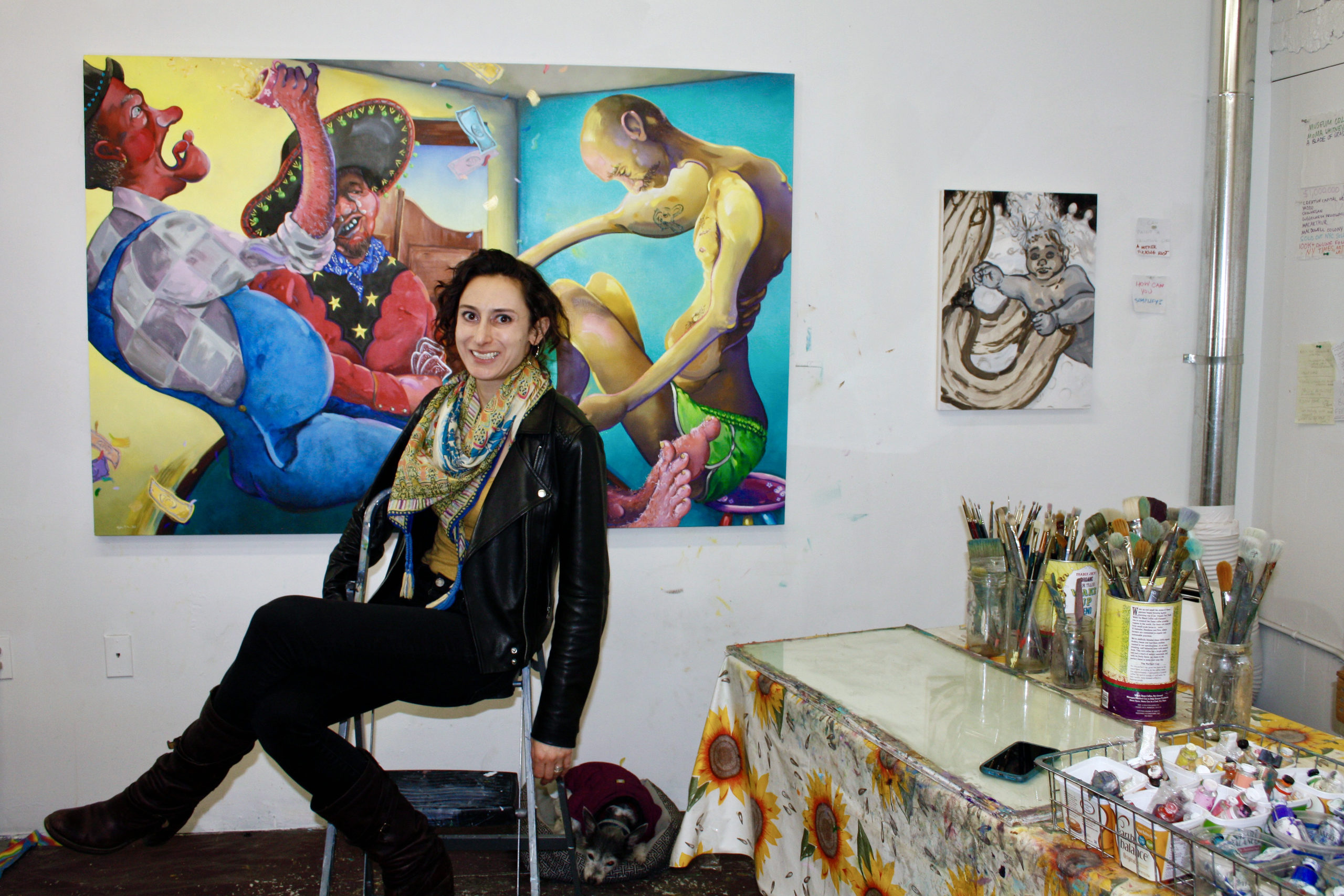 photo of Samantha Fein in front of a colorful painting with art supplies, a table and a smaller painting also in view