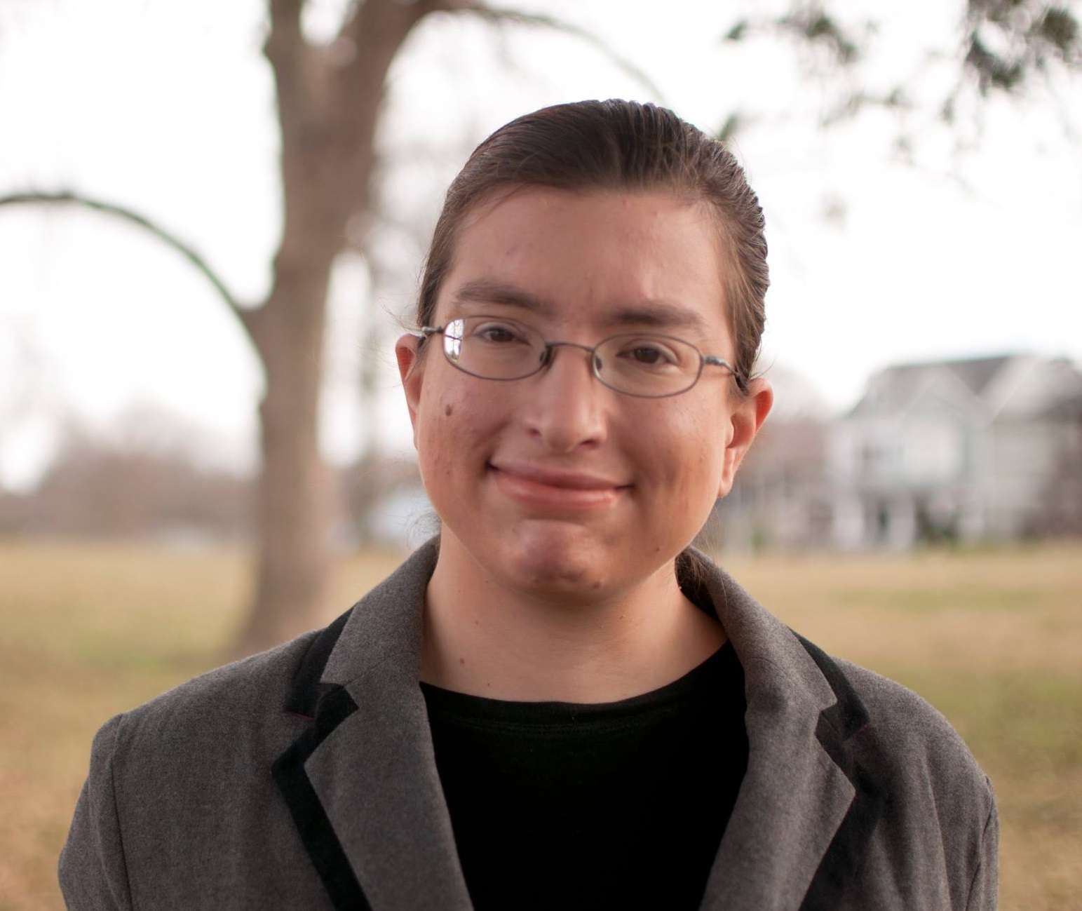 headshot of Shayna Shawn Adelman outside with a blurred image of a tree and houses in the distance