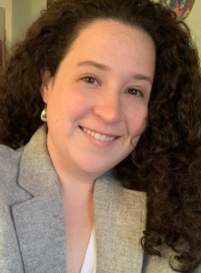 Headshot of Caitlin Brazner, white woman, curly brown hair to shoulders, smiling, grey blazer with silver drop-earrings.