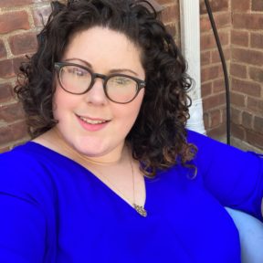 Headshot of white woman with shoulder-length curly brown hair with brown round glasses smiling with open mouth. Wearing a bright blue blouse with a gold necklace, seated in front of a brick wall. 