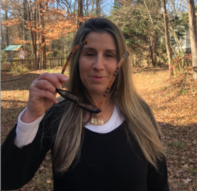 Head shot of Gisel Baler, long hair and holding glasses, wearing white and black outfit, standing in the backyard of our home in NC. 