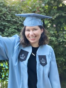 photo of Noa Rubin, a brown shoulder length haired short white woman in a blue graduation cap and gown, smiling with teeth