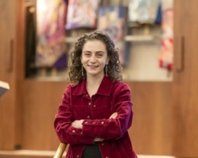  headshot of Sarina, a white woman with brown curly hair and a red corduroy jacket with arms crossed smiles at the camera with Torahs in the background