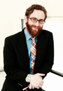 professional photo of Will Guyster at the Massachusetts Institute of Technology, white man in a jacket and tie, short reddish hair and beard, glasses, leaning on a stairwell. 