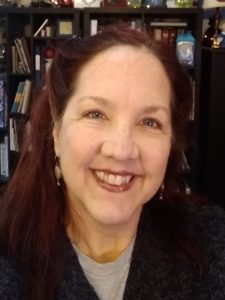 Headshot of Edie smiling, a white woman with blue eyes, red hair and wearing a grey shirt with sweater