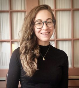 Headshot of Heather Shore; a smiling white woman wearing a black turtleneck, large glasses, and a silver hamsa necklace. She has long curly brown hair and is sitting in front of pink window panes