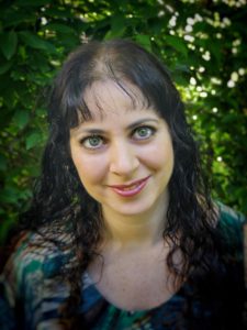 Long haired curly and freckly smiling woman in front of a lovely green tree.