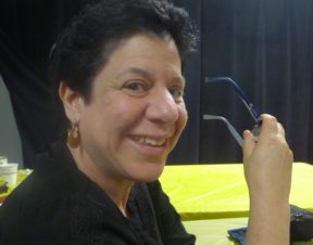 Headshot of Leah smiling, holding her glasses, wearing gold earrings; short black hair and hazel eyes.