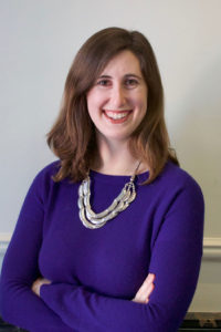 Headshot of white woman with in blue sweater and a silver necklace. Smiling with arms crossed and shoulder length brown hair