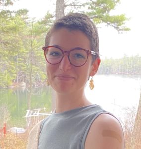  Maia has light brown cropped hair, chestnut tortoiseshell glasses, a septum ring, and gold face-shaped earrings. She stands in front of a forested lake and pale sky, wearing a moss green sleeveless shirt.