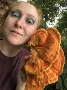  A close-up of a white, redhead, pierced-up queer in galactic drag makeup smirking and holding up a bright orange chicken of the woods mushroom with their dirty hand. 