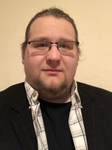 Headshot of Thomas Moser. A white male with brown hair and a goatee who is wearing glasses, a black undershirt covered by an unbuttoned white plaid collared shirt, and a suit jacket smiles at the camera. 