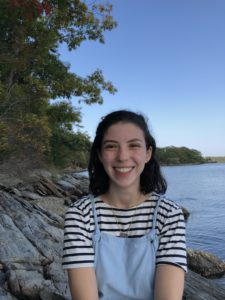 White woman with medium length brown hair wearing blue overalls smiles at the camera in front of the ocean and trees