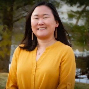  headshot of Jess Greenblatt Seeley, a Korean woman wearing a yellow blouse and gold earrings smiling