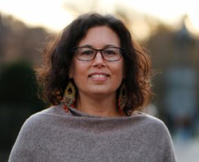 Image Description: photo of Karla Van Praag, a white woman smiling, wearing colorful glasses with shoulder-length dark brown wavy hair.