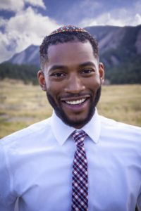 A headshot of Bryant Heinzelman, a Black man with a mustache and beard wearing a kippah, button up and tie smiling outside with a view of mountains behind