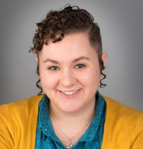A headshot of Kat Macias, a light-skin Latin person with short curly brown hair and peyos wearing a teal button up, mustard cardigan, and Star of David necklace