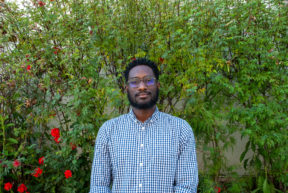 Headshot of Yehudah Webster wearing a blue button down shirt in front of a green foliage. 