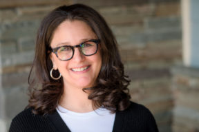 Stefanie Rhodes Headshot, woman wearing glasses and loop earrings. Wearing a white shirt with black sweater coverup.