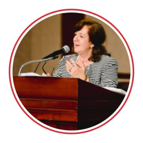 Jeannie Appleman, a white woman with brown shoulder length hair, wearing a striped shirt, standing at a podium, speaking into a microphone.