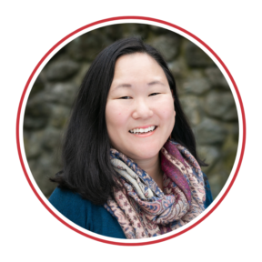 Headshot of Jess Greenblatt Seeley, a Korean woman wearing a blue top with a multi-colored scarf,smiling