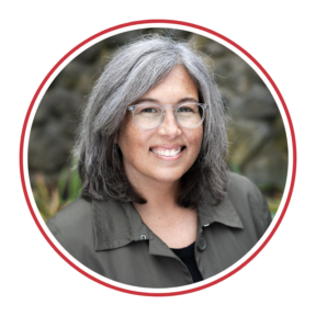 Alt Text for Karla: Headshot of Karla Van Praag, a white woman, smiling wearing clear rimmed glasses, a gray button down shirt over a black top with shoulder length hair in front of a gray stone background.